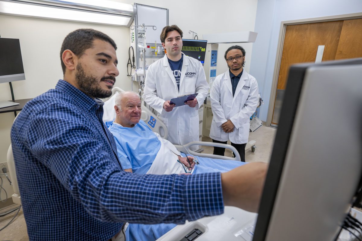 male faculty member teaching two male students in a hospital room with a patient in a bed